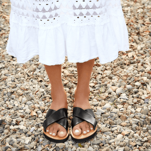 Female model wearing a white midi length skirt with the Brave Soles Constanza leather slide sandal that is sustainably made and has upcycled tire soles. Pictured in black and natural color.