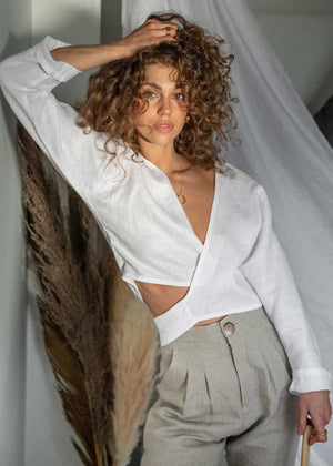 Our stunning model wearing the Avalon Tie-Up as a wrap top styled with our classic Ballina Trousers in Oat linen. She stands with her hand in her curly brown hair with pampas grass behind her.