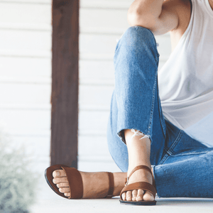 Female model in jeans and sitting down wearing the Aventura Leather walking sandal sustainably made by Brave Soles in caramel color